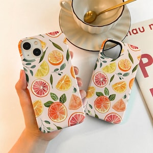 May include: Two phone cases with a white background, featuring a watercolor pattern of citrus fruits like oranges, grapefruits, and lemons, along with green leaves. The cases are held in a person's hand.