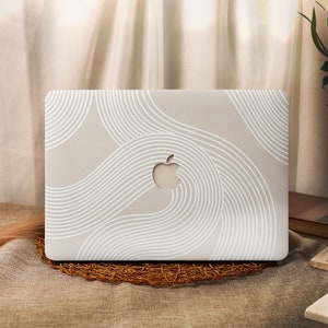 Puede incluir: Una funda para portátil beige con un diseño abstracto blanco de líneas onduladas. La funda tiene el logotipo de Apple en el centro.