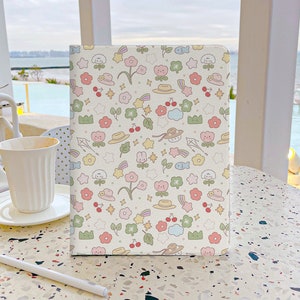 May include: A white notebook with a colorful pattern of flowers, stars, rainbows, hats, and other cute illustrations. The notebook is sitting on a table with a speckled surface.