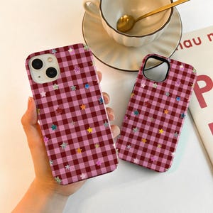Könnte beinhalten: Zwei Handyhüllen mit einem rosa-burgunderfarbenen Vichy-Muster und bunten Sternakzenten. Die Hüllen haben eine glänzende Oberfläche und sind für ein Smartphone konzipiert. Die Hüllen werden in der Hand einer Person gehalten.