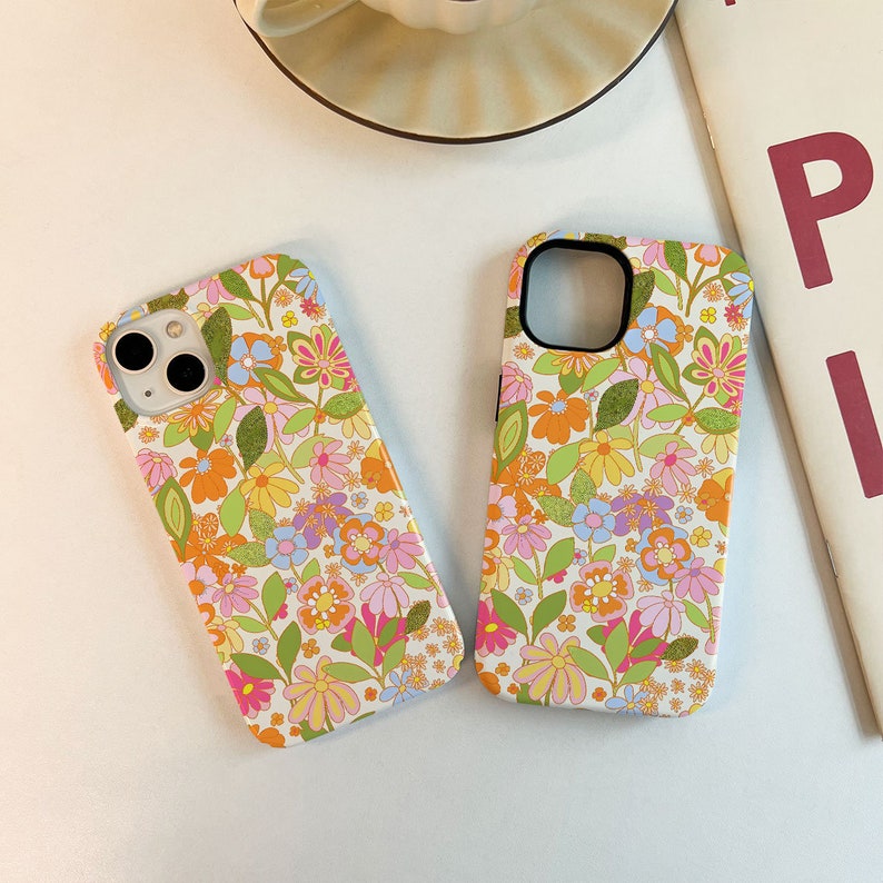 May include: Two phone cases featuring a floral design. The cases are white with a variety of colourful flowers and leaves. The cases are designed to fit an iPhone.