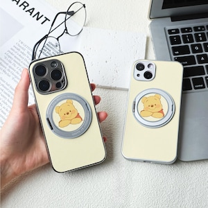May include: Two cream-coloured phone cases featuring a Winnie the Pooh character design. The cases have a silver ring holder with a clear plastic cover. The cases are on a white surface.
