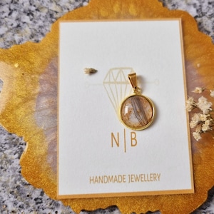 Könnte beinhalten: Ein goldfarbener Anhänger mit einem runden, transparenten Mittelteil, der Haarsträhnen und Goldflocken enthält. Der Anhänger liegt auf einer weißen Karte mit den Initialen "N B" und den Worten "HANDMADE JEWELLERY". Die Karte liegt auf einer goldfarbenen Oberfläche.