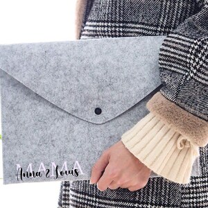 Könnte beinhalten: Eine hellgraue Filz-Dokumententasche mit schwarzem Knopfverschluss. Der Text "MAMA Anna & Louis" ist in rosa und schwarzer Schrift auf der Vorderseite aufgedruckt. Die Tasche wird von einer Person gehalten, die einen schwarz-weiß karierten Mantel trägt.