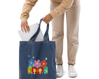 May include: A denim tote bag with a colorful floral design and two figures embracing within a heart. The bag is being held by a person, and a white laptop is placed inside. The bag has a blue denim color.