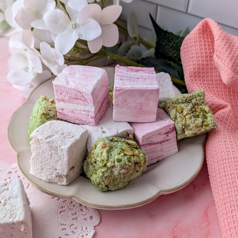 May include: A plate of assorted marshmallows, including pink and white flavored marshmallows and green marshmallows with pistachio pieces.
