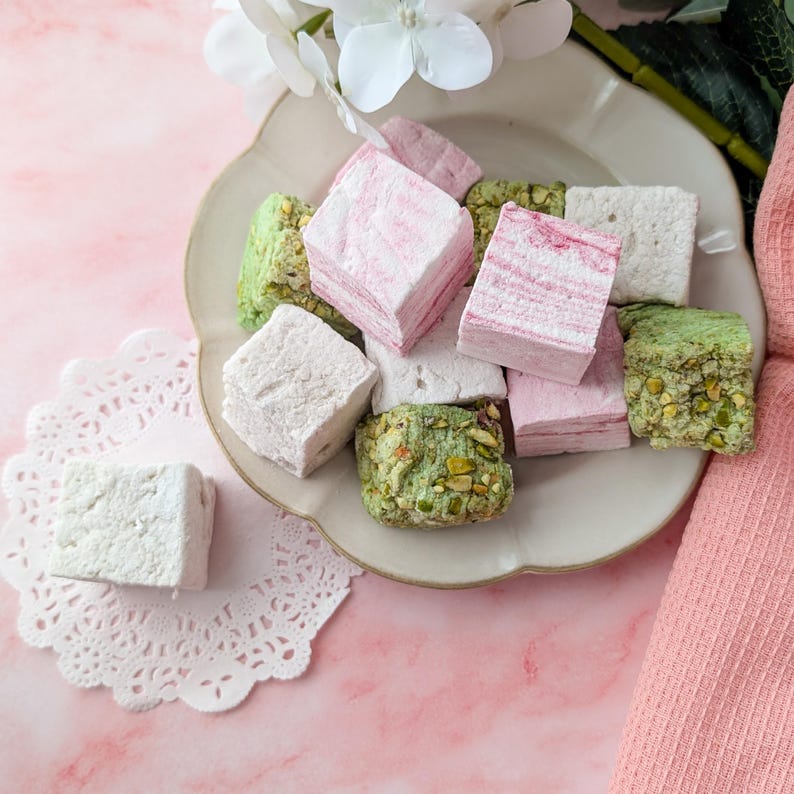 May include: A plate of pink, green, and white marshmallow squares. The marshmallows are arranged in a random pattern on a white plate with a scalloped edge.