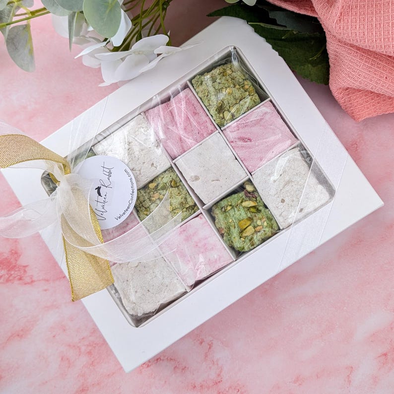 May include: A white box with a clear lid filled with nine squares of pink and green marshmallow. The box is tied with a gold ribbon and a white label with the text "Natalie Rabbit" and "Valentines Collection".