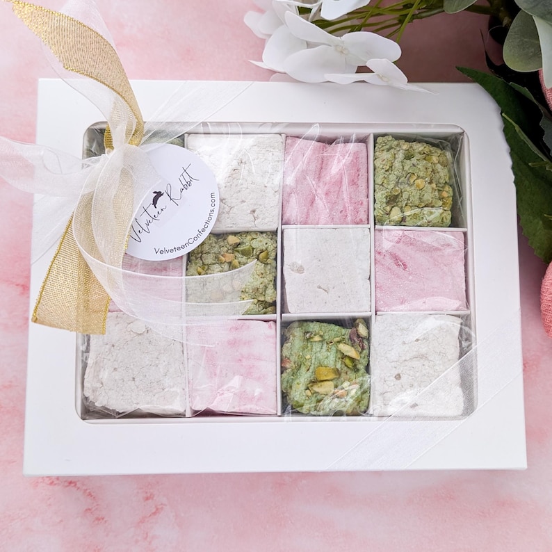 May include: A white gift box with a clear lid filled with nine squares of pastel pink and green marshmallow squares. The box is tied with a gold ribbon and a white tag with the text "Velveteen Rabbit VelveteenConfections.com".