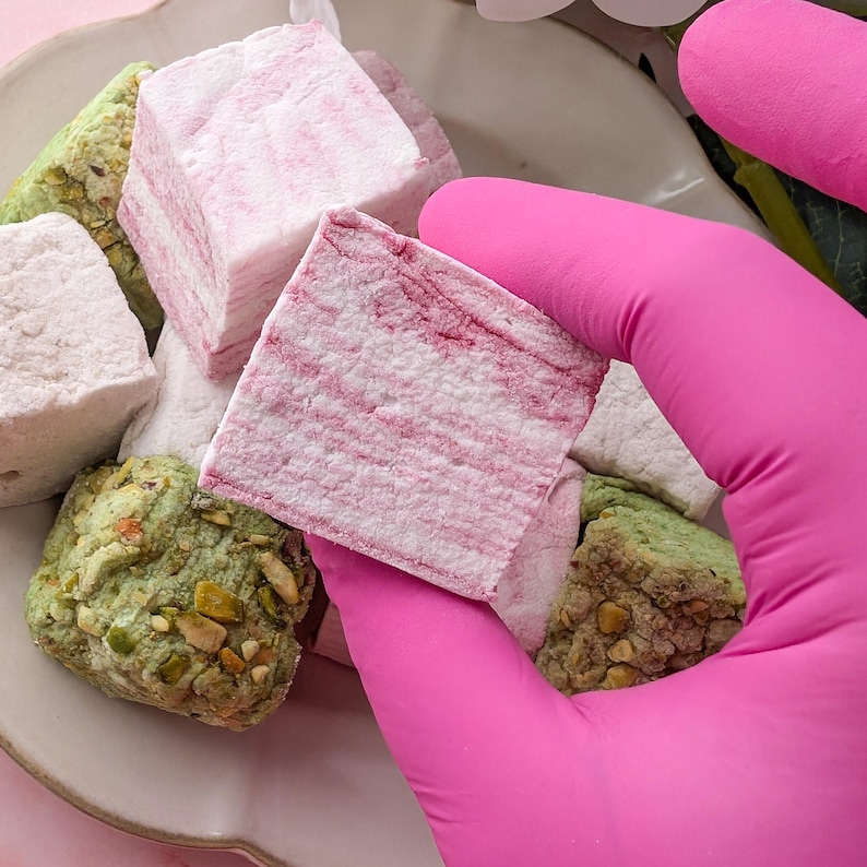 May include: A close-up of a pink-gloved hand holding a pink and white striped marshmallow. Other marshmallows in various colors and flavors are visible on a white plate.