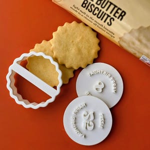 May include: A close-up of golden-brown butter biscuits with a scalloped edge, a white cookie cutter, and two white stamps. The stamps read "Happy Birthday" and feature a number. The biscuit packaging reads "Butter Biscuits".