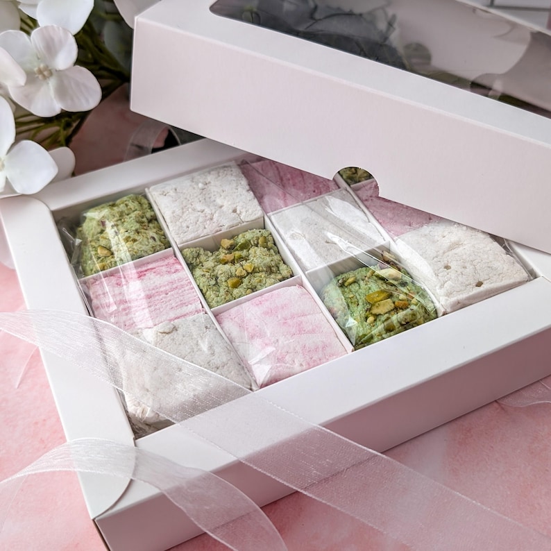 May include: A white box with a clear lid filled with small squares of pink, white, and green marshmallow. The box is tied with a white ribbon.
