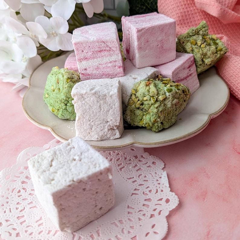 May include: A plate of assorted marshmallows, including pink, white, and green varieties. The marshmallows are dusted with powdered sugar and some are topped with chopped pistachios.
