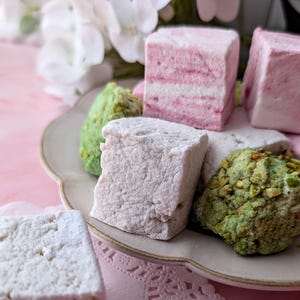 May include: A plate of assorted marshmallows, including pink, white, and green varieties. The marshmallows are arranged on a white plate with a scalloped edge.