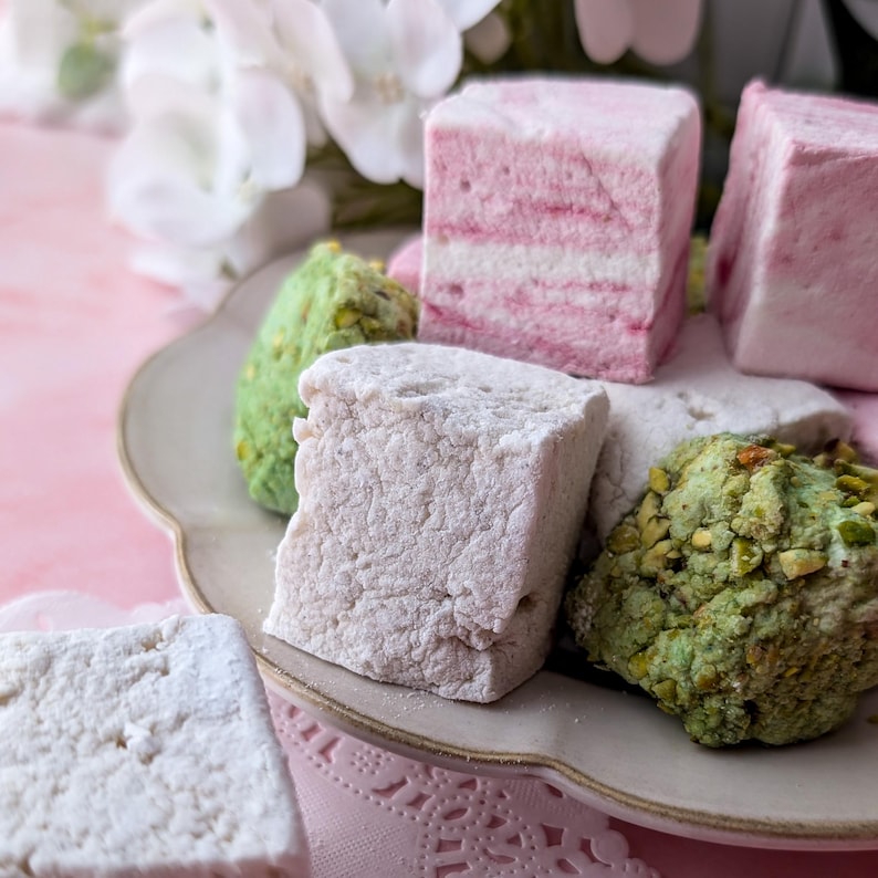 May include: A plate of assorted marshmallows, including pink, white, and green varieties. The marshmallows are arranged on a white plate with a scalloped edge.