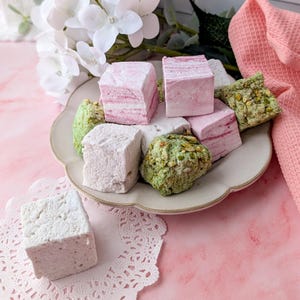May include: A plate of assorted marshmallows, including pink, white, and green flavors. The marshmallows are arranged in a pile on a white plate with a scalloped edge. The plate is sitting on a pink surface.