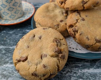 May include: A close-up of a stack of chocolate chip cookies on a blue plate. The cookies are golden brown and have a slightly crispy texture. The cookies are arranged in a pile, with the largest cookie on the bottom and the smallest cookie on top.