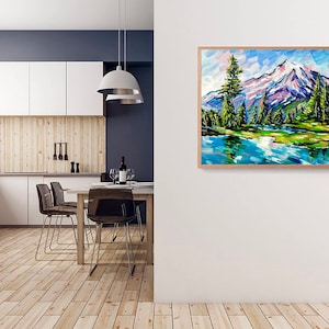 May include: A framed landscape painting depicts a mountain scene with a snow-capped peak, green trees, and a blue lake. The artwork uses bold brushstrokes and vibrant colors, creating a textured effect. The frame is made of light-colored wood.