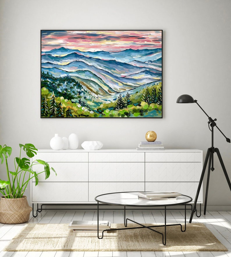 May include: A painting of a mountain landscape with a pink and blue sky. The mountains are painted in shades of blue, green, and brown. The painting is framed in black.