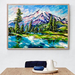 May include: A framed landscape painting depicts a snow-capped mountain range reflected in a blue lake. The artwork features vibrant brushstrokes in shades of blue, green, pink, and white, with tall green trees lining the water's edge. The painting is in a light brown wooden frame.