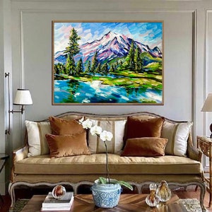 May include: An original landscape painting featuring a snow-capped mountain, a serene lake, and lush green trees. The artwork is framed in a natural wood tone and hangs above a sofa with brown and white pillows. The painting's style is impressionistic.