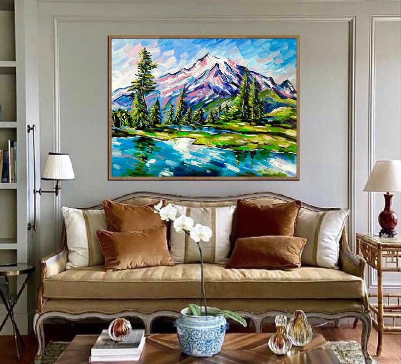 May include: An original landscape painting featuring a snow-capped mountain, a serene lake, and lush green trees. The artwork is framed in a natural wood tone and hangs above a sofa with brown and white pillows. The painting's style is impressionistic.