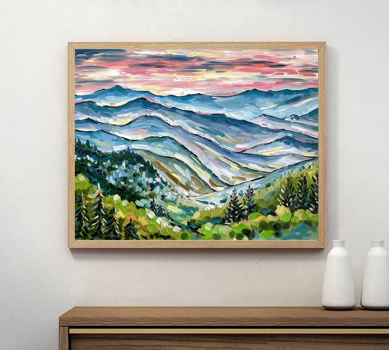 May include: A painting of a mountain landscape with a pink and blue sky. The mountains are painted in shades of blue, green, and yellow. The painting is framed in a light wood frame.