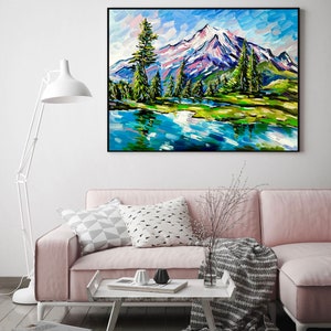 May include: An original oil painting of a mountain landscape with a lake. The artwork features a snow-capped mountain, green trees, and a blue lake reflecting the sky. The painting is framed in a black frame and hangs above a pink sofa.