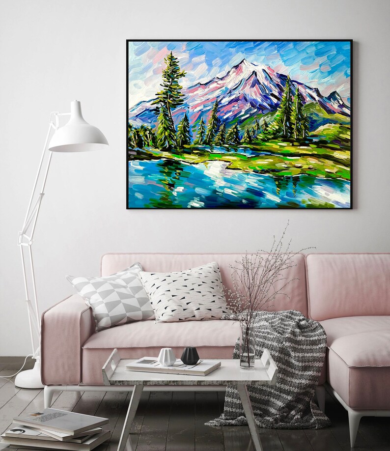 May include: An original oil painting of a mountain landscape with a lake. The artwork features a snow-capped mountain, green trees, and a blue lake reflecting the sky. The painting is framed in a black frame and hangs above a pink sofa.