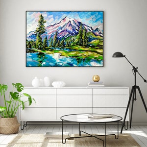 May include: A framed landscape painting depicts a mountain range with snow-capped peaks, a serene lake, and evergreen trees. The artwork features vibrant brushstrokes in shades of blue, green, and white, creating a textured effect. The painting is displayed above a white cabinet.