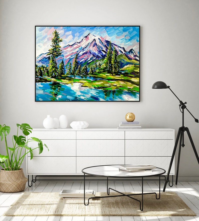 May include: A framed landscape painting depicts a mountain range with snow-capped peaks, a serene lake, and evergreen trees. The artwork features vibrant brushstrokes in shades of blue, green, and white, creating a textured effect. The painting is displayed above a white cabinet.
