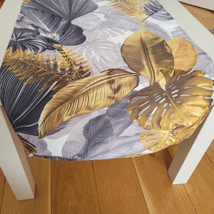May include: A table runner with a botanical design. It features gold and grey leaves on a white background. The runner is placed on a white table, showcasing the leaf pattern.