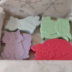 May include: Four colorful cat-shaped soaps in a white box. The soaps are lavender, light blue, green, and red.