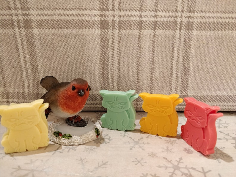 May include: Five small, grumpy cat-shaped soaps in different colors: yellow, green, orange, and pink. The soaps are arranged in a row on a white surface with a small robin figurine in the background.