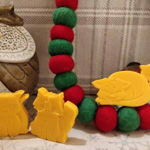 May include: Four yellow cat-shaped soap bars. Two cats are sitting upright, one is lying down, and one is standing with its tail curled around its body. The soap bars are on a background of red and green felt balls.