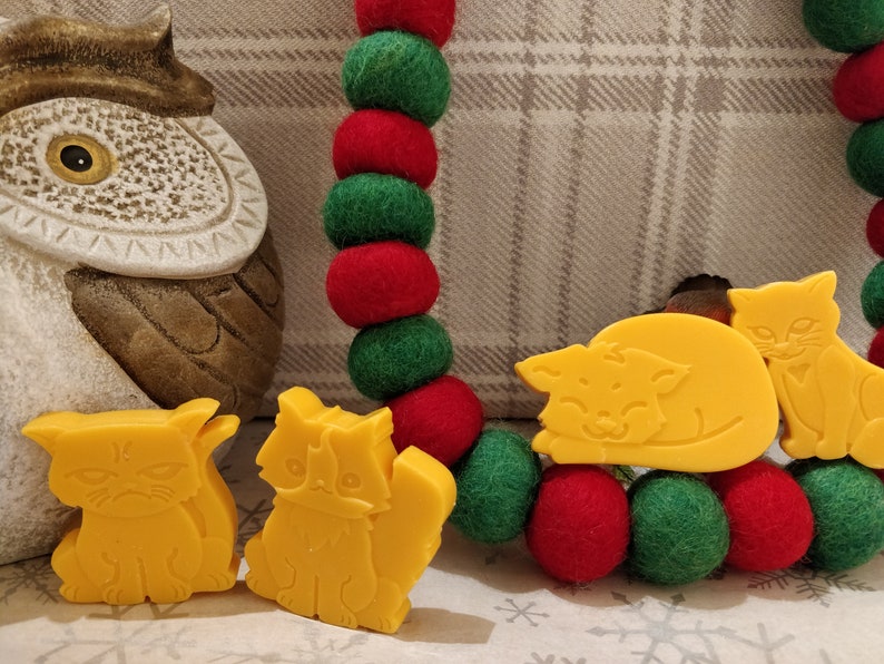 May include: Four yellow cat-shaped soap bars. Two cats are sitting upright, one is lying down, and one is standing with its tail curled around its body. The soap bars are on a background of red and green felt balls.