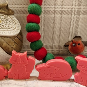 May include: A set of four pink cat-shaped soap bars with different expressions. The soap bars are arranged in a row on a white surface with a red and green garland and a brown owl figurine in the background.