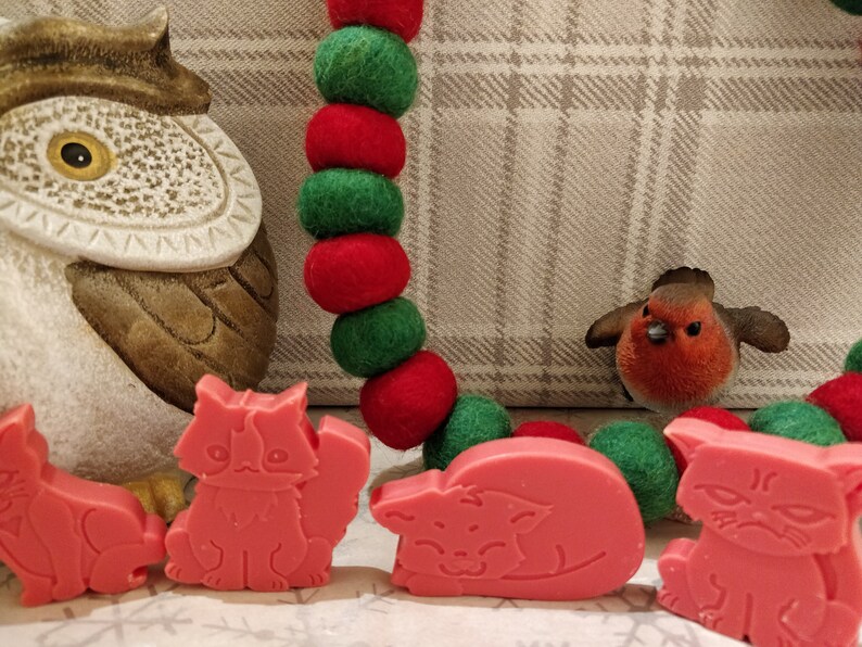 May include: A set of four pink cat-shaped soap bars with different expressions. The soap bars are arranged in a row on a white surface with a red and green garland and a brown owl figurine in the background.