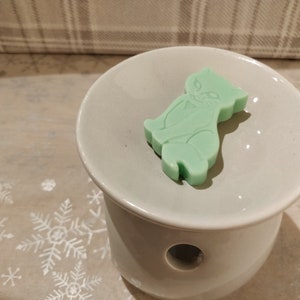 May include: A light green wax melt shaped like a cat sitting on a white ceramic wax warmer.