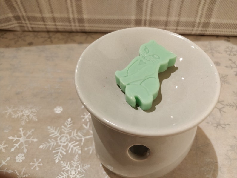 May include: A light green wax melt shaped like a cat sitting on a white ceramic wax warmer.