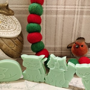 May include: Four green cat-shaped soap bars. The cats are in different poses, including sleeping, sitting, and standing. The soap bars are on a white surface with a red and green garland in the background.