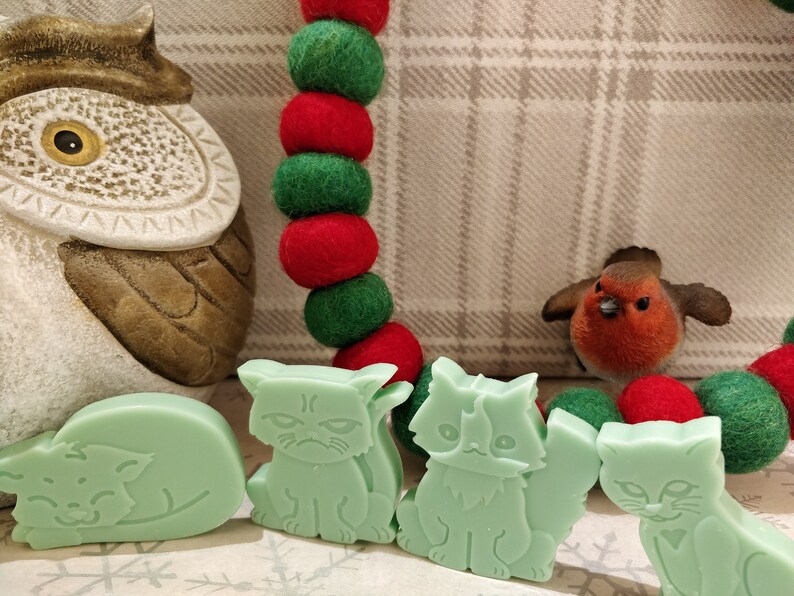 May include: Four green cat-shaped soap bars. The cats are in different poses, including sleeping, sitting, and standing. The soap bars are on a white surface with a red and green garland in the background.