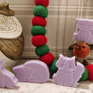 May include: Four lavender-colored cat-shaped soap bars, two sitting upright, one lying down, and one standing on a small red robin figurine. The soaps are on a background of a plaid fabric and a garland of red and green felt balls.