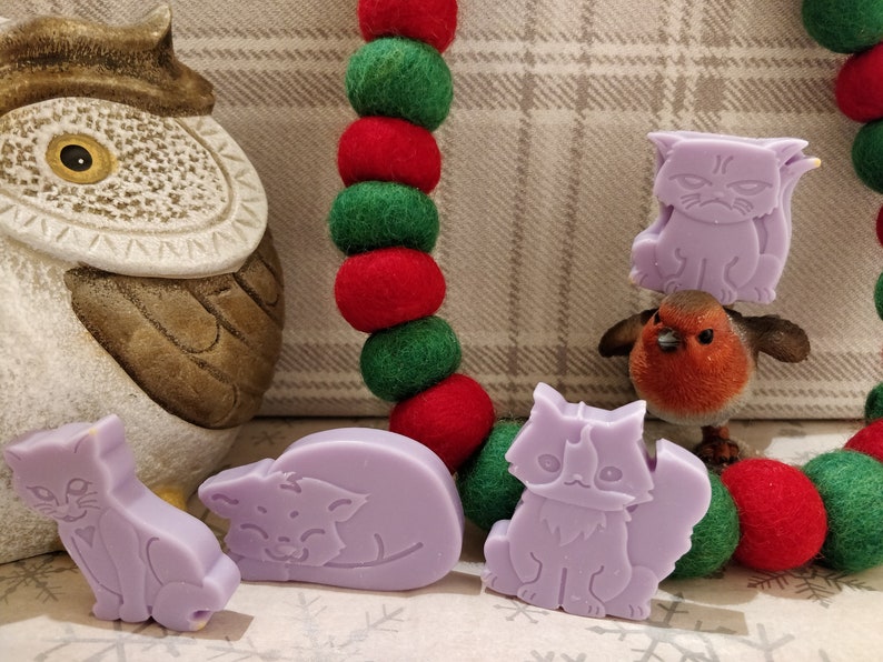 May include: Four lavender-colored cat-shaped soap bars, two sitting upright, one lying down, and one standing on a small red robin figurine. The soaps are on a background of a plaid fabric and a garland of red and green felt balls.