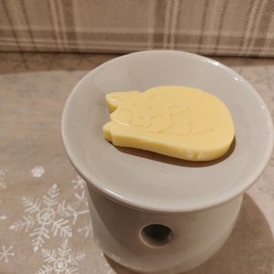 May include: A white ceramic wax warmer with a round top and a hole in the center. A yellow wax melt shaped like a sleeping cat is on top of the warmer.