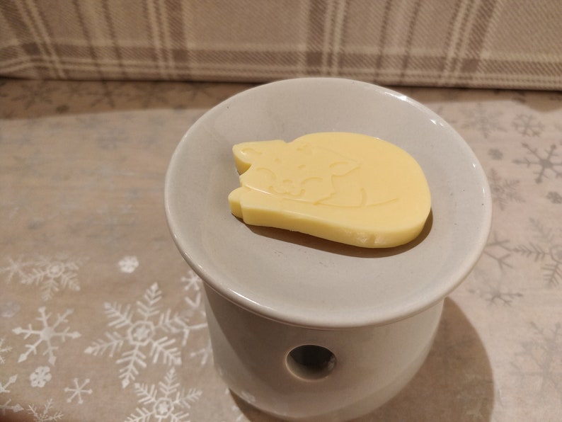 May include: A white ceramic wax warmer with a round top and a hole in the center. A yellow wax melt shaped like a sleeping cat is on top of the warmer.