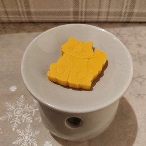 May include: A yellow wax melt shaped like a cat sitting on a white ceramic wax warmer.