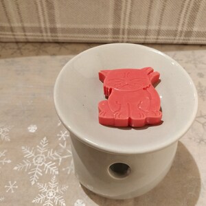 May include: A red wax melt shaped like a cat sitting on a white ceramic wax warmer.