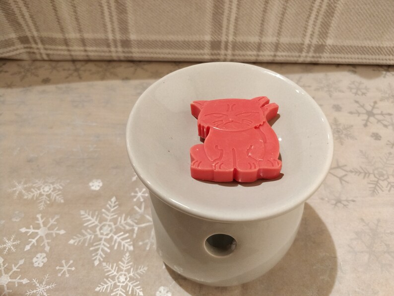 May include: A red wax melt shaped like a cat sitting on a white ceramic wax warmer.