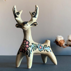 Puede incluir: Una figurita de cerámica blanca de ciervo con diseños florales negros y verdes y flores rosas y azules. El ciervo tiene grandes cuernos y una expresión caprichosa.